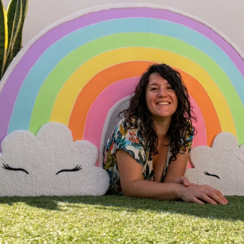 Claire Lys allongée devant un grand décor arc-en-ciel pastel à louer, sur une pelouse ensoleillée