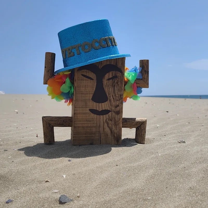 Mascotte totem en bois de Totem, chapeau bleu et colliers de fleurs, plantée sur une plage de l'Hérault face à la mer