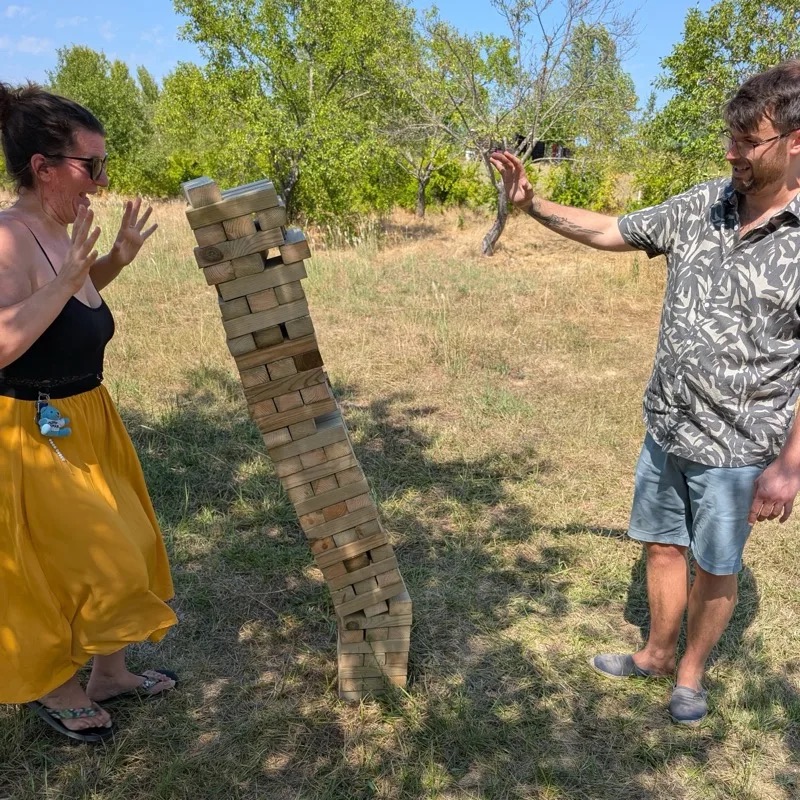 Une tour de Jenga géant penchée à l'extrême dans un jardin, entre deux joueurs qui retiennent leur souffle, les mains en l'air