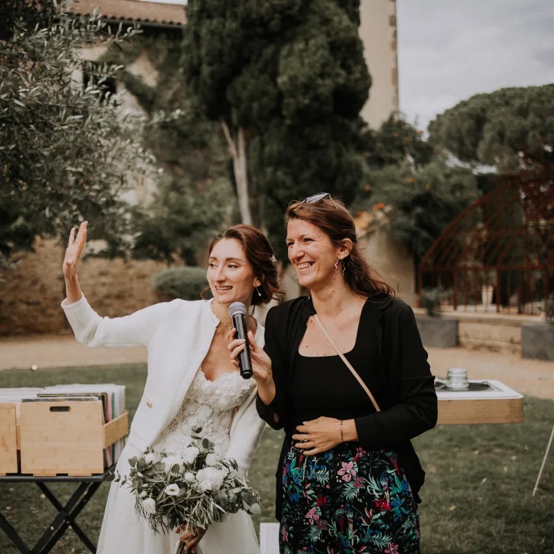 Claire Lys, micro en main, anime une cérémonie de mariage aux côtés de la mariée souriante et son bouquet, dans un jardin de domaine